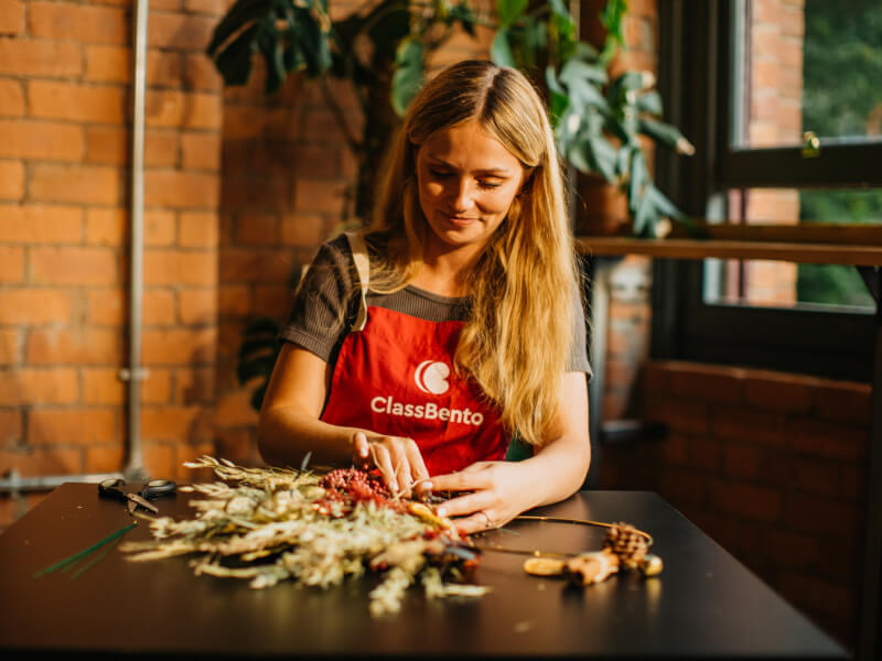 A woman is sitting at a table making a festive wreath from greenery and berries
